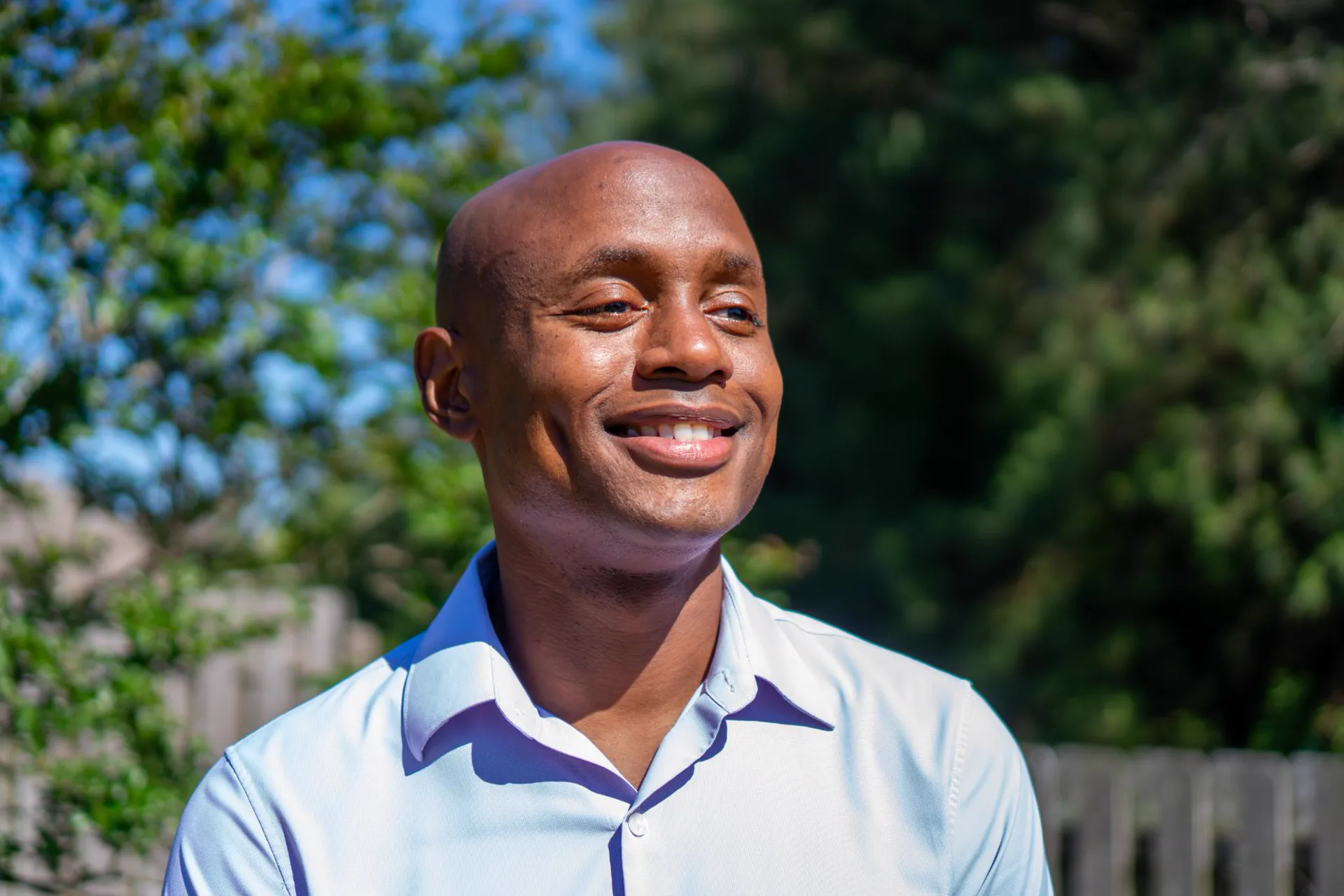 Michael C. Shedrick smiling outdoors against green trees and a clear blue sky